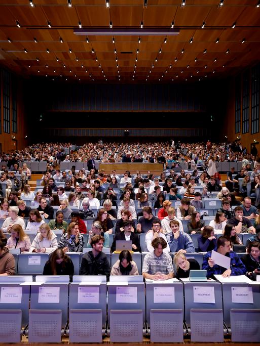 Studierende sitzen in einem großen Hörsaal. In der ersten Reihe sind noch Plätze frei, die mit Namensschildern belegt sind.