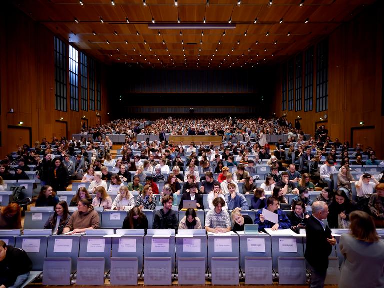 Studierende sitzen in einem großen Hörsaal. In der ersten Reihe sind noch Plätze frei, die mit Namensschildern belegt sind.