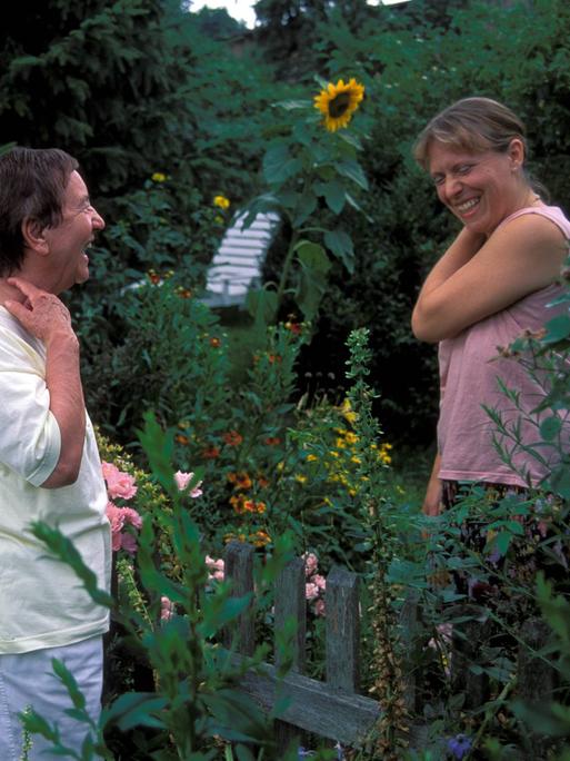 Zwei Frauen stehen beim Gespräch am Gartenzaun zwischen ihren Gärten und lachen.