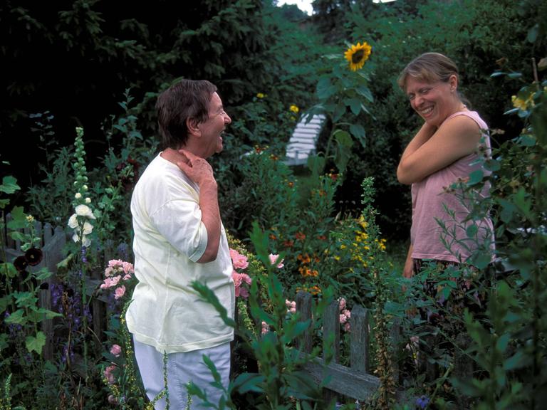 Zwei Frauen stehen beim Gespräch am Gartenzaun zwischen ihren Gärten und lachen.