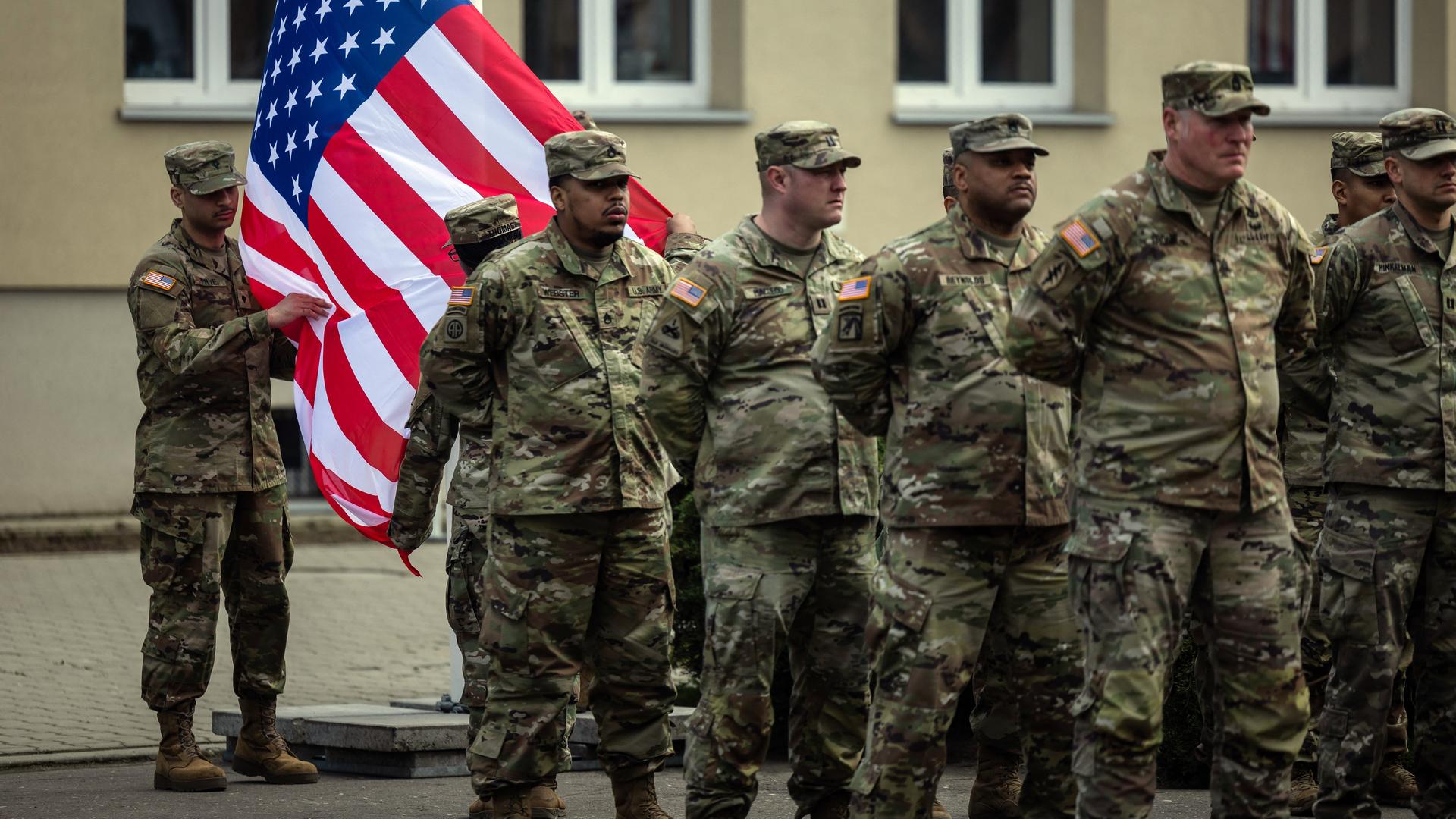 Das Foto zeigt US-Soldaten vor einer Fahne der USA. Im Hintergrund ist ein Haus zu sehen.