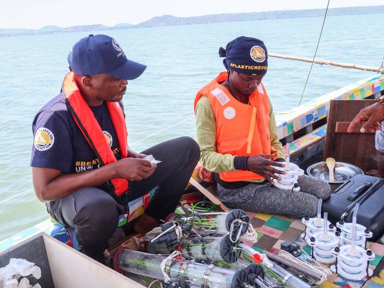 Zwei Männer sitzen auf einem Boot, vor ihnen sind Plastikdosen und -behälter, mit denen Wasserproben aus dem Victoriasee genommen werden. Der rechte Mann ist Kenias Superstar Juliani.