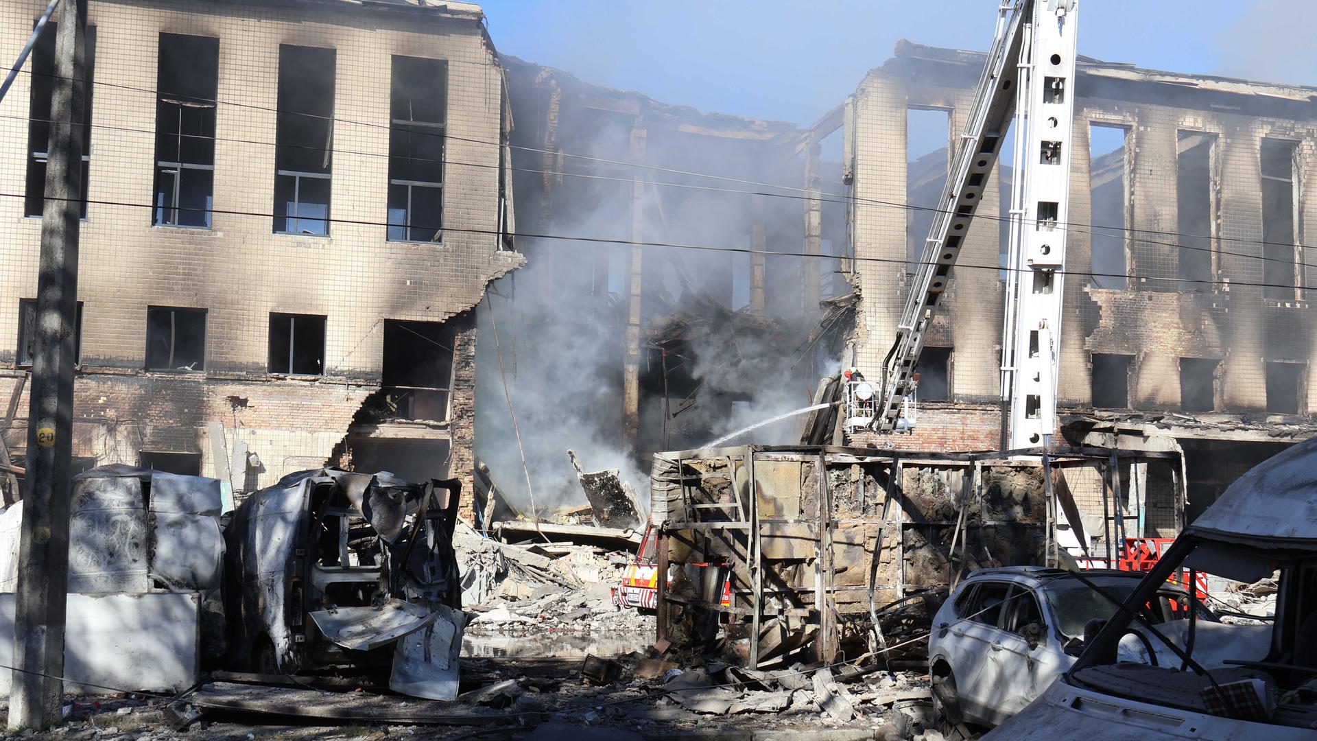 Feuerwehrleute löschen einen Brand in einem völlig zerstörten Haus, davor stehen ausgebrannte Autowracks.
