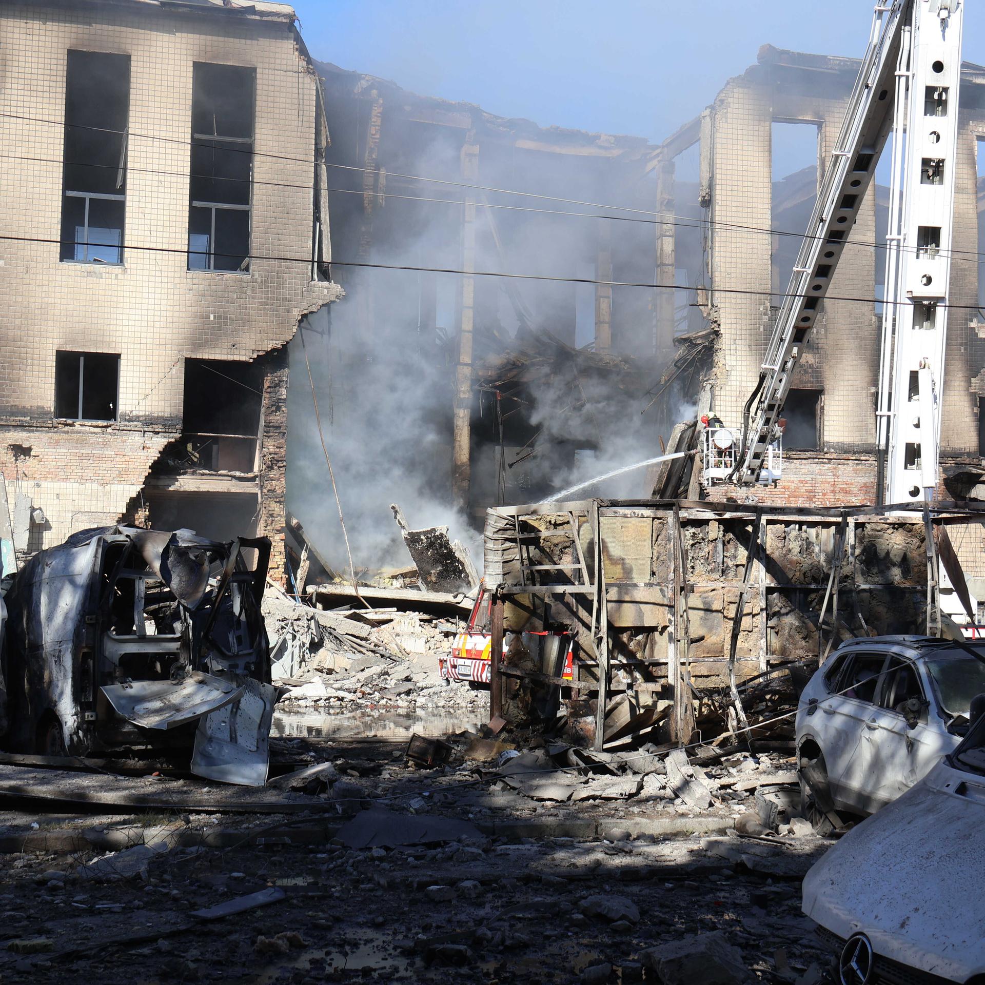 Feuerwehrleute löschen einen Brand in einem völlig zerstörten Haus, davor stehen ausgebrannte Autowracks.