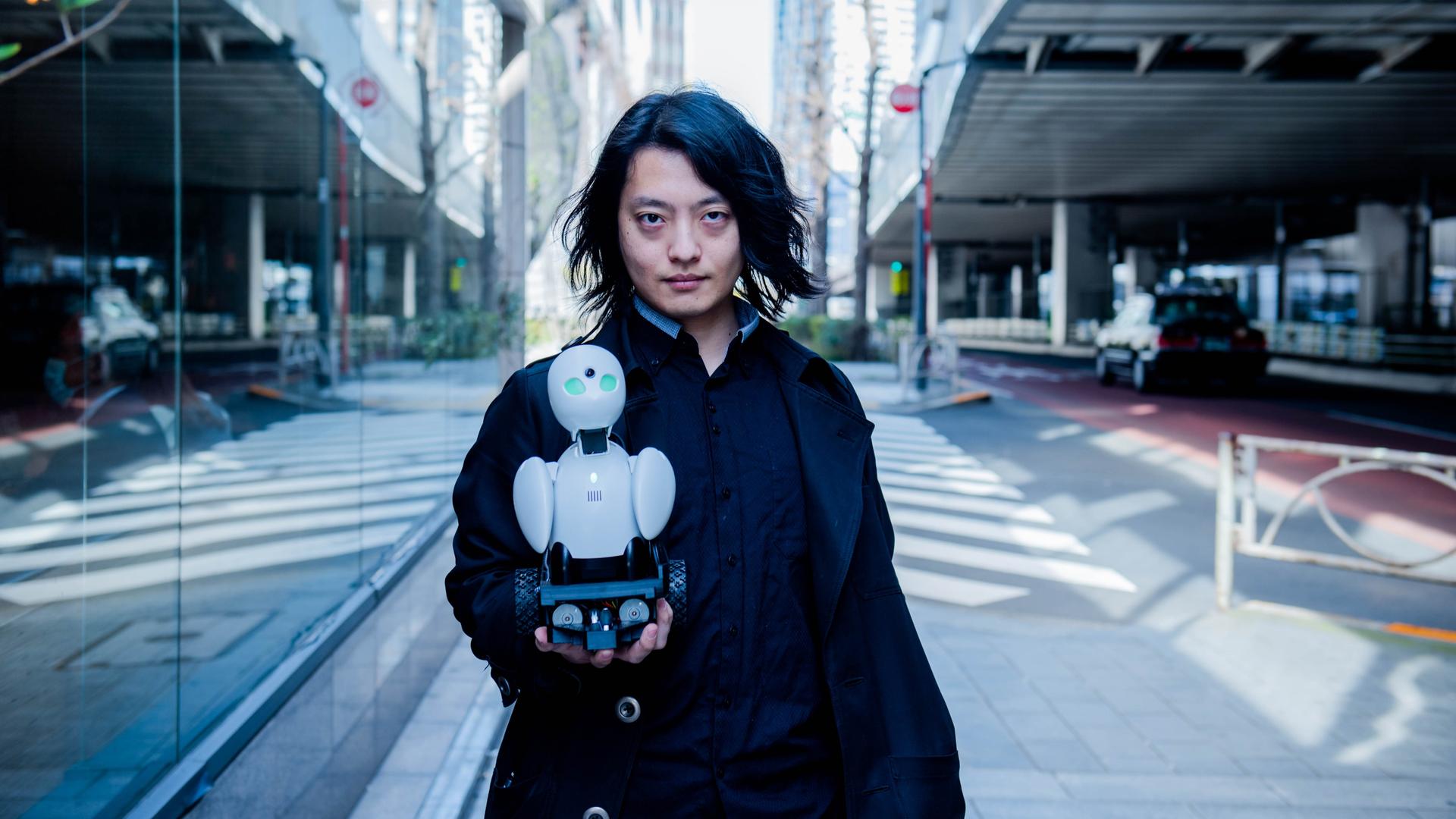 Der japanische Medienkünstler Ory Yoshifuji trägt ein weites schwarzes Sakko und hält in seiner rechten Hand einen 20 cm hohen weißen Roboter mit grünen Augen.
