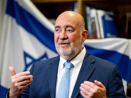 Ron Prosor, Botschafter Israels in Deutschland, steht vor einer israelischen Flagge und gestikuliert mit den Händen.