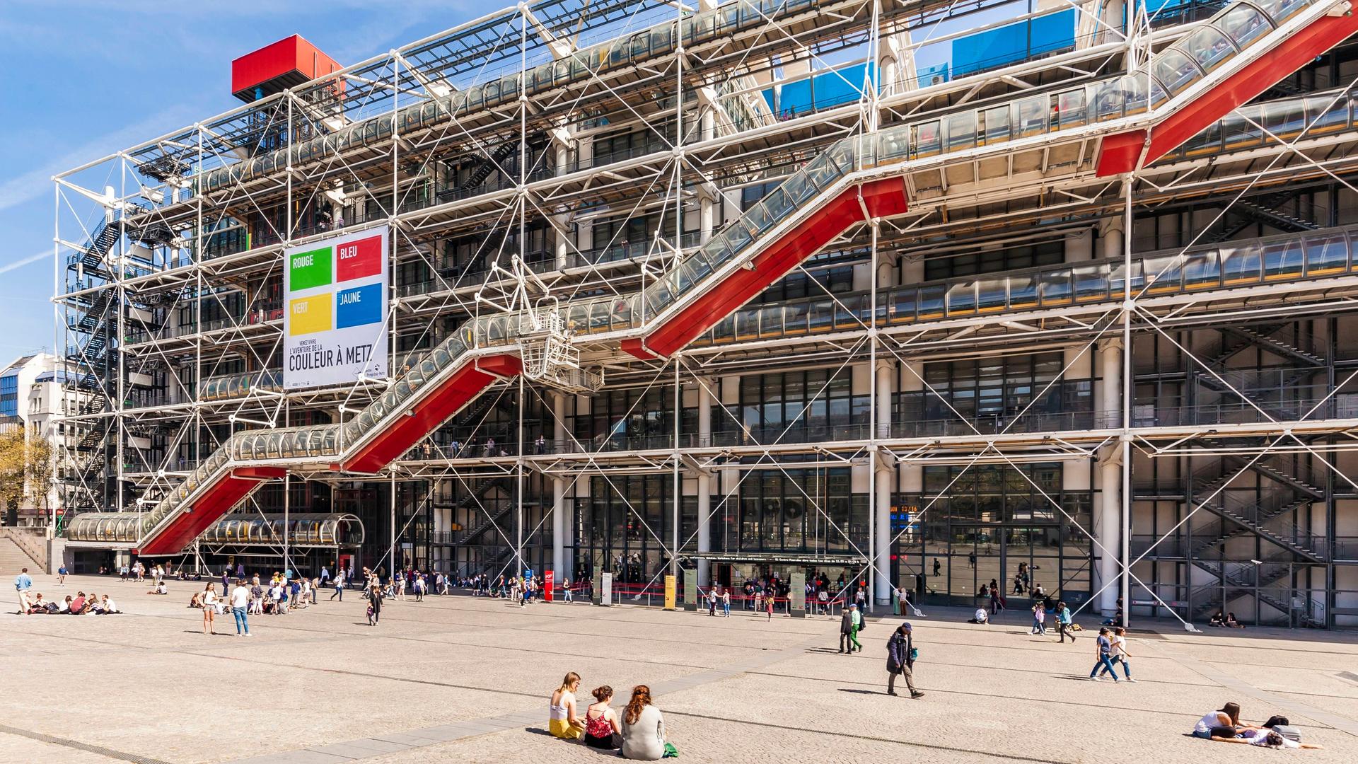 Die Fassade des Museums Centre Pompidou in Paris mit den spezifischen bunten Röhren. Auf dem Platz davor sitzen Menschen in leichter Kleidung auf dem Boden.