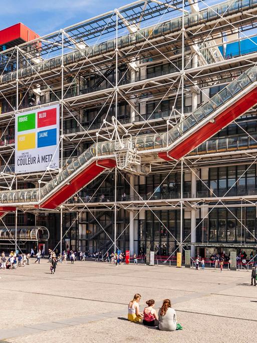 Das Centre Pompidou in Paris mit den spezifischen bunten Röhren. Auf dem Platz davor sitzen Menschen in leichter Kleidung auf dem Boden. 