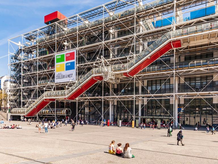Das Centre Pompidou in Paris mit den spezifischen bunten Röhren. Auf dem Platz davor sitzen Menschen in leichter Kleidung auf dem Boden. 