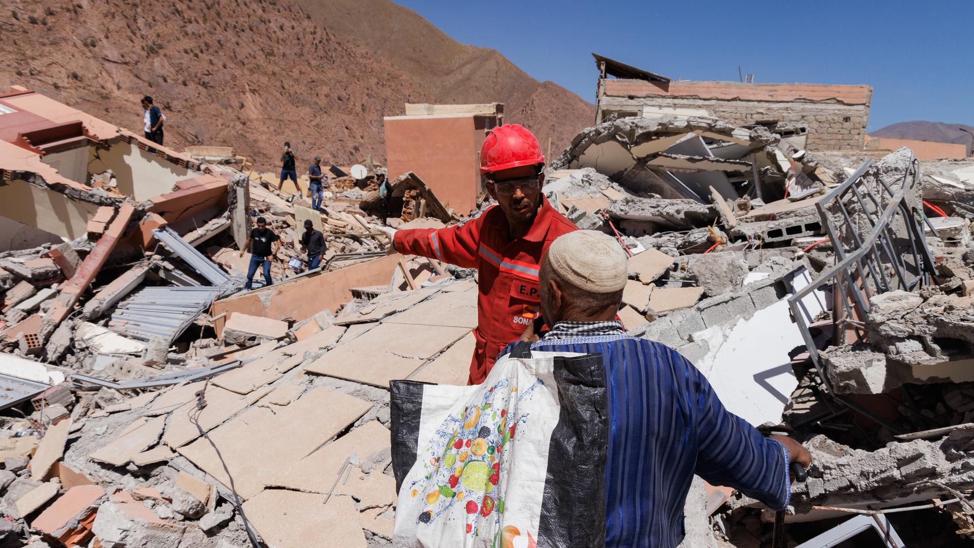 Ein Mann mit Helm und Schutzweste spricht mit einem Zivilisten inmitten völlig zerstörter Häuser vor einem Berghang.