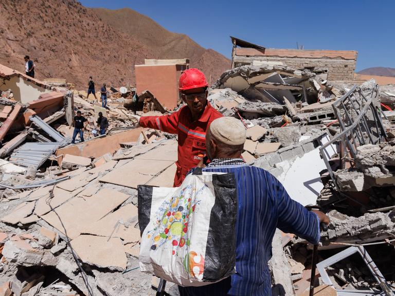 Ein Mann mit Helm und Schutzweste spricht mit einem Zivilisten inmitten völlig zerstörter Häuser vor einem Berghang.