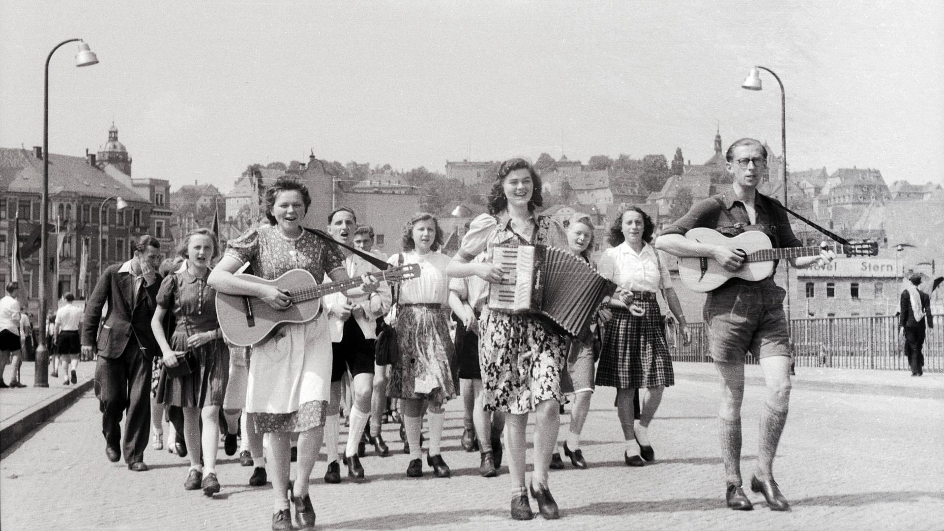 Junge Frauen und Männer laufen durch eine Straße. Die vorderen spielen dabei Gitarre und Ziehharmonika.