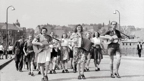 Junge Frauen und Männer laufen durch eine Straße. Die vorderen spielen dabei Gitarre und Ziehharmonika.