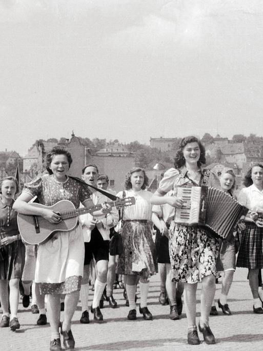 Junge Frauen und Männer laufen durch eine Straße. Die vorderen spielen dabei Gitarre und Ziehharmonika.