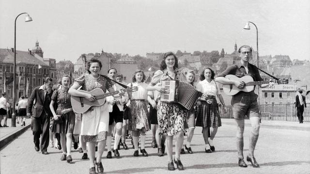 Junge Frauen und Männer laufen durch eine Straße. Die vorderen spielen dabei Gitarre und Ziehharmonika.