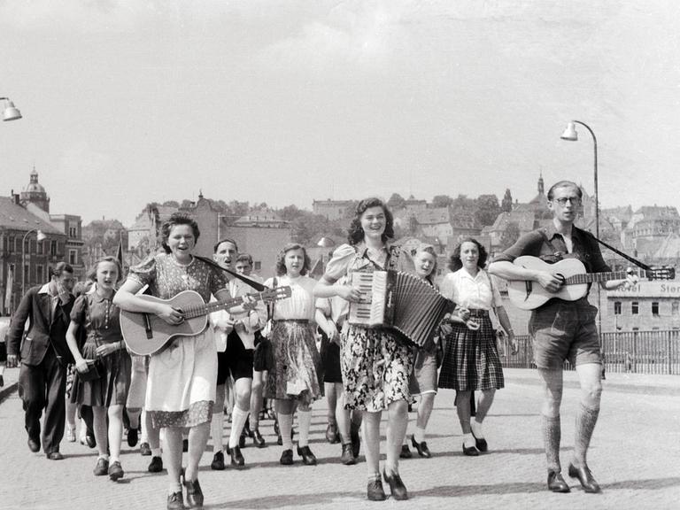 Junge Frauen und Männer laufen durch eine Straße. Die vorderen spielen dabei Gitarre und Ziehharmonika.