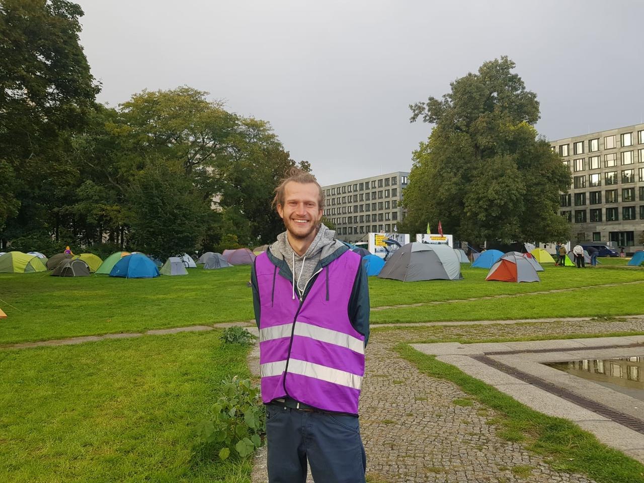 Aktivist Jan Gerber mit lila Sicherheitsweste, im Hintergrund ist ein Protestcamp zu sehen.