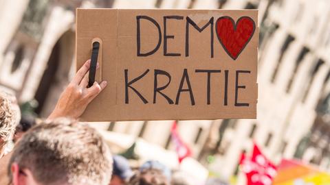 Bei einer Kundgebung wird ein Schild mit der Aufschrift "Demokratie" in die Höhe gestreckt. Dabei ist das O in Demokratie in Form eines Herzes.