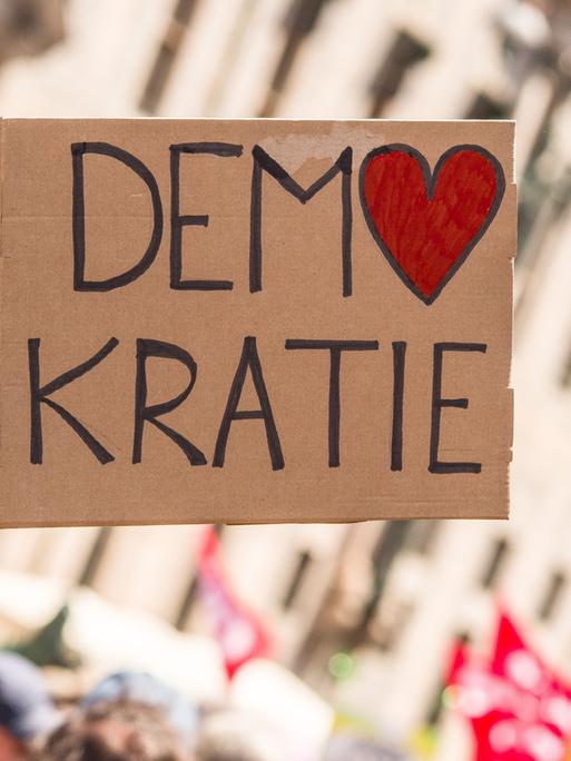 Bei einer Kundgebung wird ein Schild mit der Aufschrift "Demokratie" in die Höhe gestreckt. Dabei ist das O in Demokratie in Form eines Herzes.