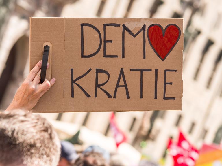 Bei einer Kundgebung wird ein Schild mit der Aufschrift "Demokratie" in die Höhe gestreckt. Dabei ist das O in Demokratie in Form eines Herzes.