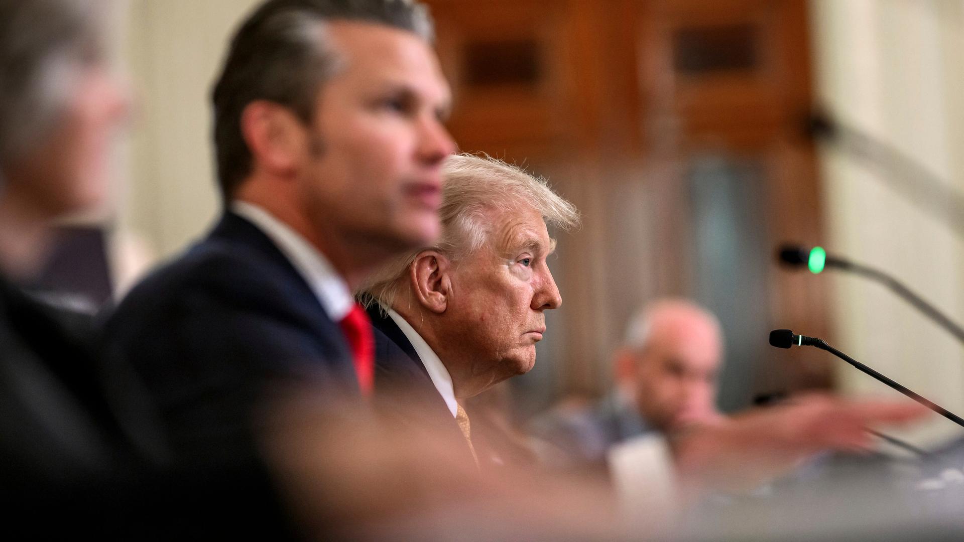 Zwei Männer sitzen an einem Tisch. Vor ihnen Mikrofone. Der jüngere Mann im Vordergrund ist unscharf. Beide blicken zum rechten Bildrand.