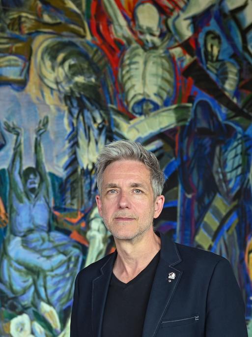 André Nicke steht im Foyer des Schwedter Theaters vor dem monumentalen Wandbild "Triumph des Todes, Triumph des Lebens" des DDR-Künstlers Ronald Paris auf dem fragmentarisch Figuren in blau, grün und gelb zu erkennen sind.