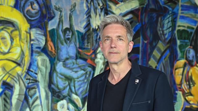 André Nicke steht im Foyer des Schwedter Theaters vor dem monumentalen Wandbild "Triumph des Todes, Triumph des Lebens" des DDR-Künstlers Ronald Paris auf dem fragmentarisch Figuren in blau, grün und gelb zu erkennen sind.