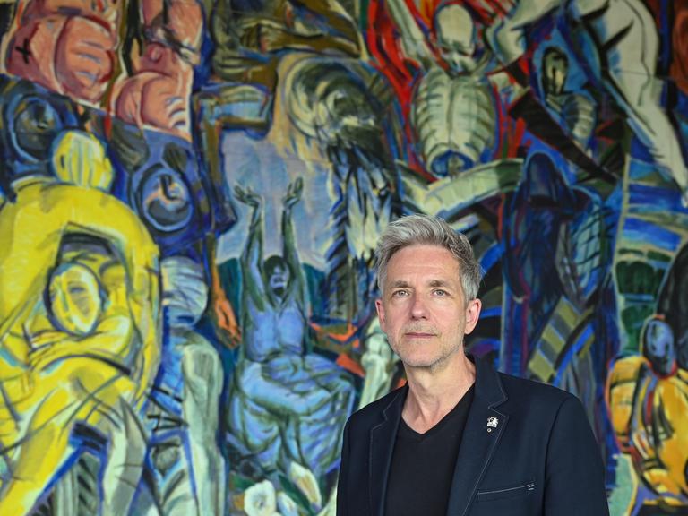 André Nicke steht im Foyer des Schwedter Theaters vor dem monumentalen Wandbild "Triumph des Todes, Triumph des Lebens" des DDR-Künstlers Ronald Paris auf dem fragmentarisch Figuren in blau, grün und gelb zu erkennen sind.