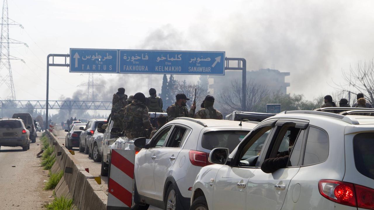 In einem Verkehrsstau Richtung Qardaha, Syrien, sind bewaffnete Kämpfer zu erkennen, die sich aus dem Autofenster lehnen. Im Hintergrund sind Rauchschwaden zu erkennen. Sie stammen mutmaßlich von den Kämpfen im Westen Syriens.  