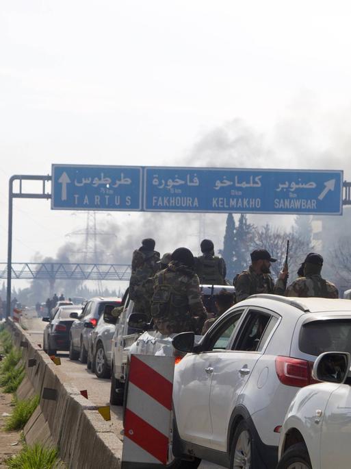 In einem Verkehrsstau Richtung Qardaha, Syrien, sind bewaffnete Kämpfer zu erkennen, die sich aus dem Autofenster lehnen. Im Hintergrund sind Rauchschwaden zu erkennen. Sie stammen mutmaßlich von den Kämpfen im Westen Syriens.  