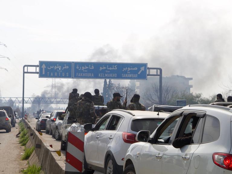 In einem Verkehrsstau Richtung Qardaha, Syrien, sind bewaffnete Kämpfer zu erkennen, die sich aus dem Autofenster lehnen. Im Hintergrund sind Rauchschwaden zu erkennen. Sie stammen mutmaßlich von den Kämpfen im Westen Syriens.  