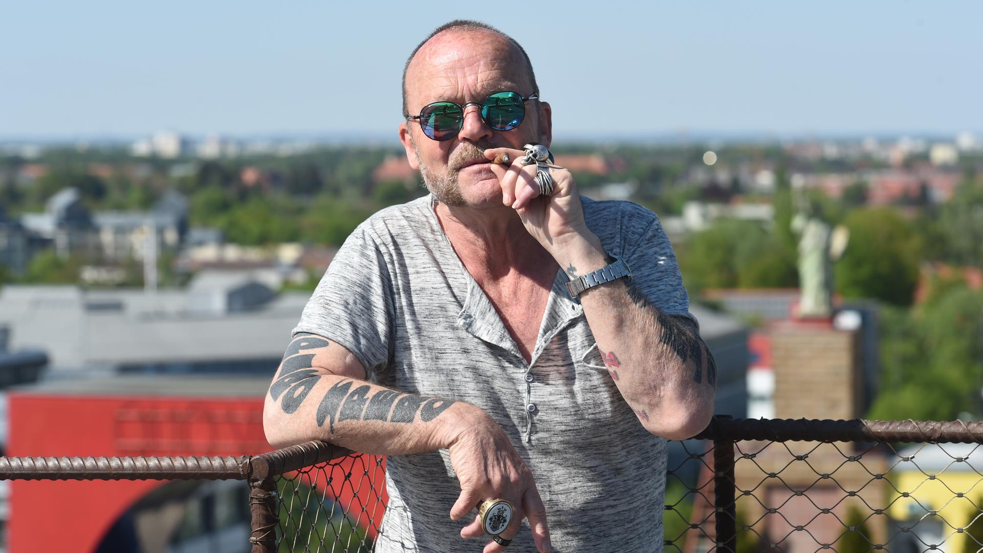 Ein tätowierter Mann mit Sonnenbrille steht eine Zigarre rauchend auf einem Hausdach. Ein tätowierter Mann mit Sonnenbrille steht eine Zigarre rauchend auf einem Hausdach.