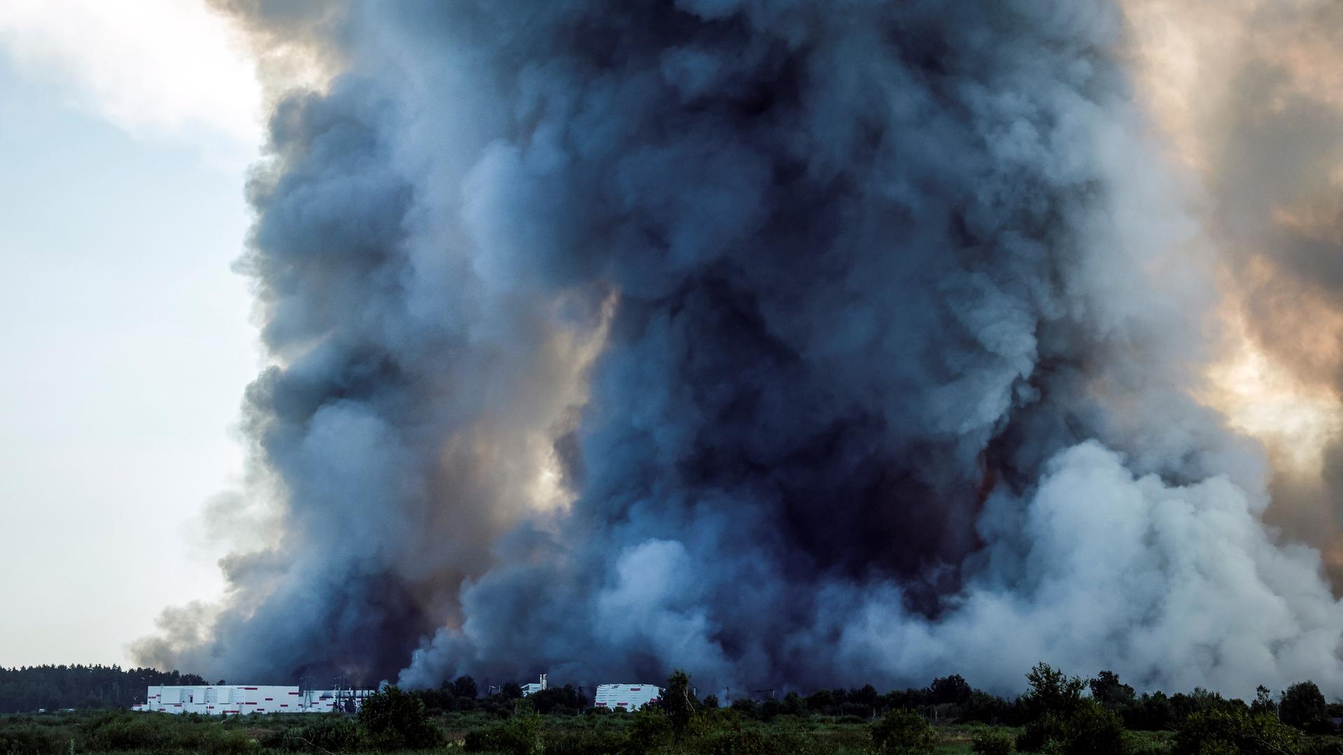 Das Bild zeigt eine große Rauch-Wolke. Die Rauch-Wolke steigt in den Himmel. Unten im Bild ist eine Lager-Halle zu sehen. Das Bild zeigt eine große Rauch-Wolke. Die Rauch-Wolke steigt in den Himmel. Unten im Bild ist eine Lager-Halle zu sehen.