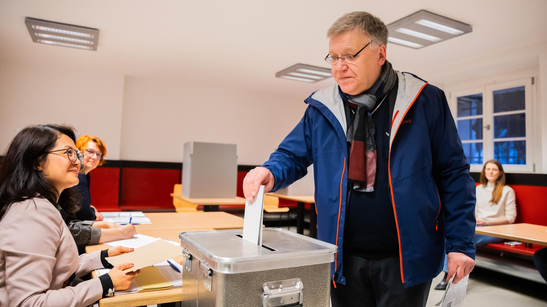 Ein Mann wirft einen Wahlzettel in eine Wahlurne. Zwei Frauen an einem Tisch davor lächeln ihn an.