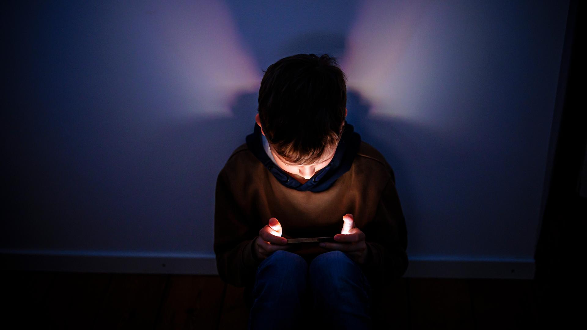Symbolfoto. Gestellte Aufnahme zum Thema Kinder nutzen Social Media. Ein Junge sitzt in einem dunklen Raum und wird von einem Smartphone beleuchtet. 
