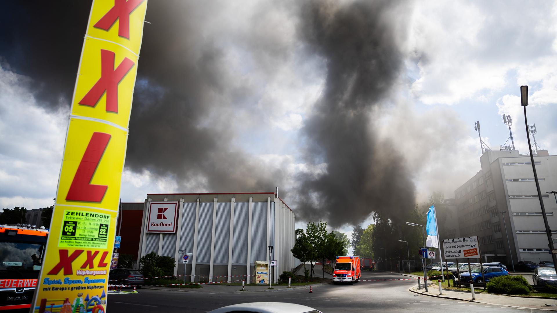 Blick aus einiger Entfernung: Auf einer Straße steht ein Rettungswagen neben einer "Kaufland"-Filiale, Hinter dieser steig dichter schwarzer Qualm in den bewölkten Himmel. Am linken vorderen Bildrand ein senkrechtes Werbebanner an einer Laterne mit der Aufschrift "XXL".