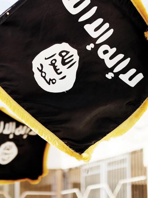 Im Vordergrund halten zwei Hände die schwarze Flagge der Terrormiliz „Islamischer Staat“ (IS). Im Hintergrund ist eine weitere solche Flagge zu sehen. 