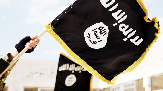 Im Vordergrund halten zwei Hände die schwarze Flagge der Terrormiliz „Islamischer Staat“ (IS). Im Hintergrund ist eine weitere solche Flagge zu sehen. 