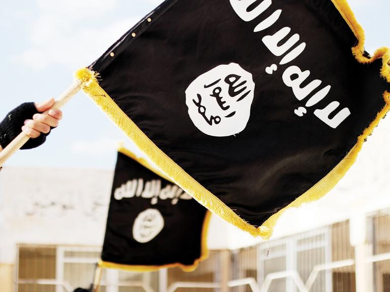 Im Vordergrund halten zwei Hände die schwarze Flagge der Terrormiliz „Islamischer Staat“ (IS). Im Hintergrund ist eine weitere solche Flagge zu sehen. 