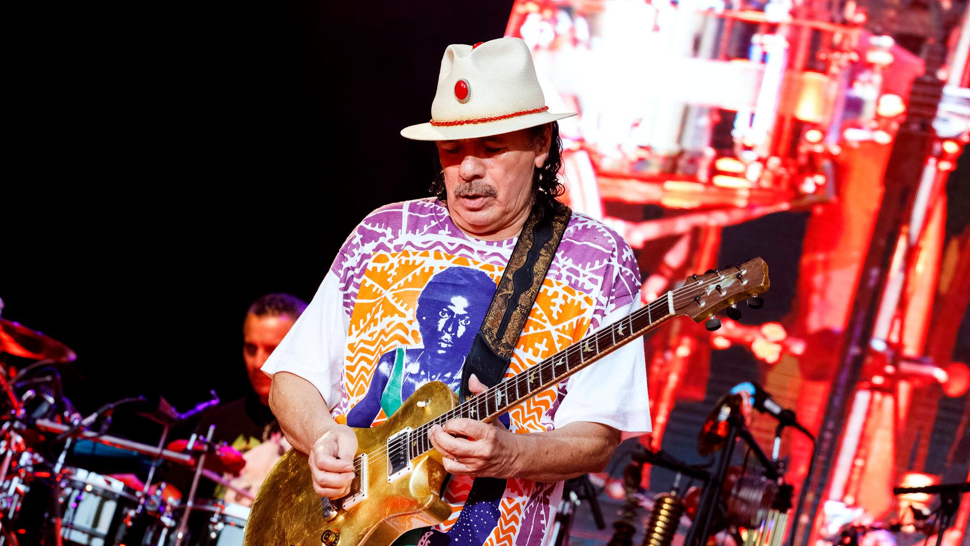 Carlos Santana steht auf der Bühne und spielt Gitarre. Er trägt einen weißen Hut und ein quietschbuntes, weites Oberteil. 