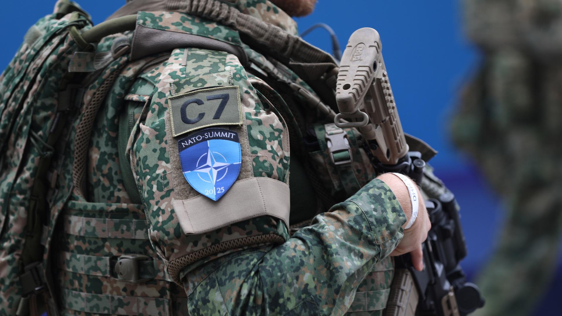Eine bewaffnete Person in olivfarbener Tarnkleidung. Auf dem Ärme der Uniform ist das blaue NATO-Emblem genäht. Eine bewaffnete Person in olivfarbener Tarnkleidung. Auf dem Ärme der Uniform ist das blaue NATO-Emblem genäht.