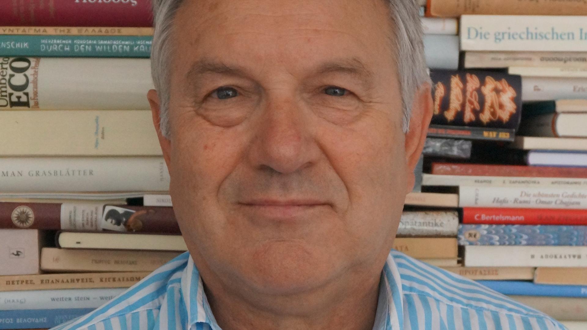 Der Dichter und Kommunalpolitiker Costas Gianacacos steht vor einer Wand aus Büchern.