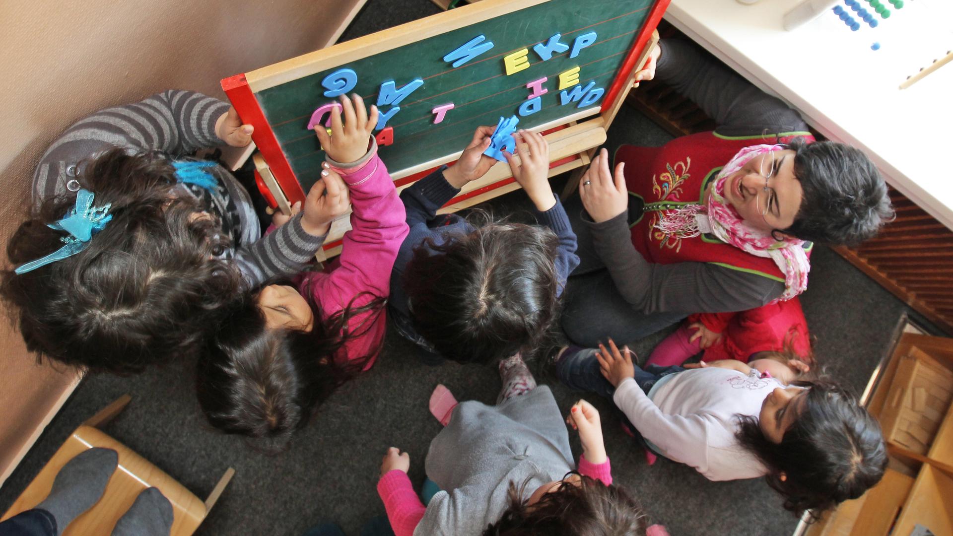 Blick von oben auf eine Schulklasse. Die türkische Erzieherin nutzt Buchstaben mit denen die Kinder an einer Tafel Worte bilden. 