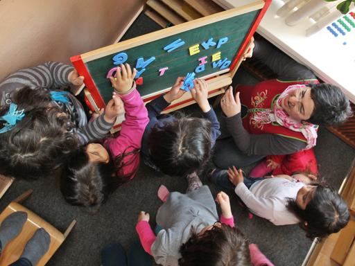 Blick von oben auf eine Schulklasse. Die türkische Erzieherin nutzt Buchstaben mit denen die Kinder an einer Tafel Worte bilden. 