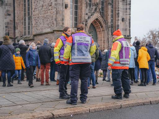 Einsatzkräfte in orangefarbenen Jacken und andere Menschen stehen vor der Trauerstelle am Dom nach dem Anschlag auf den Magdeburger Weihnachtsmarkt