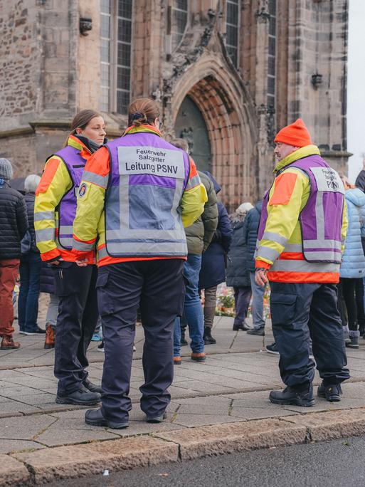 Einsatzkräfte in orangefarbenen Jacken und andere Menschen stehen vor der Trauerstelle am Dom nach dem Anschlag auf den Magdeburger Weihnachtsmarkt