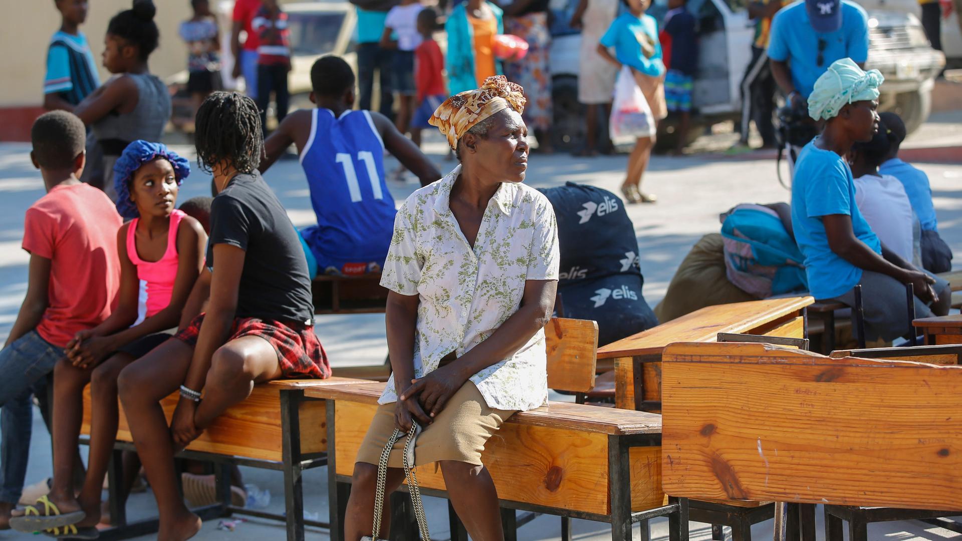 Mehrere Menschen sitzen vor einer Schule, die im Bezirk der Brothers Road in Petion-Ville als Unterkunft dient, auf Möbeln und warten. 