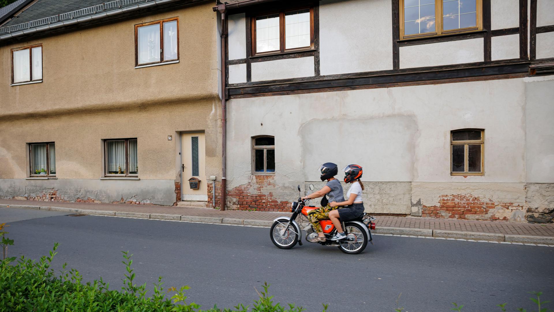 Ein Junge und ein Mädchen fahren auf einem Simson Moped an alten Häusern vorbei über eine Dorfstrasse.