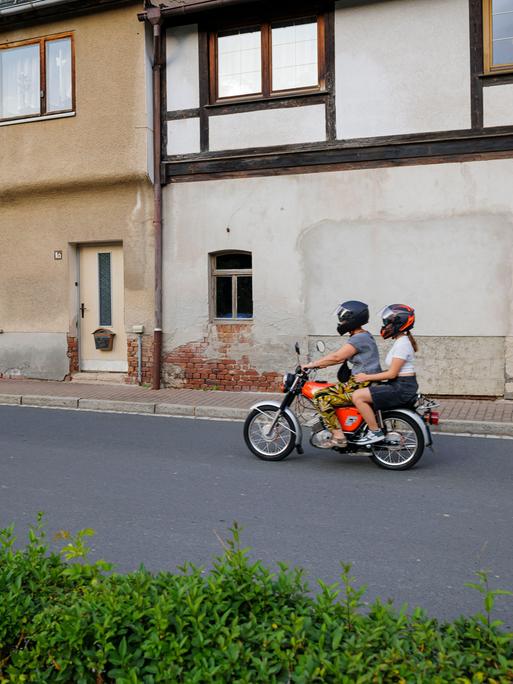 Ein Junge und ein Mädchen fahren auf einem Simson Moped an alten Häusern vorbei über eine Dorfstrasse.