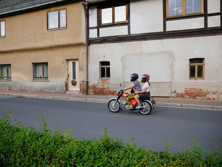 Ein Junge und ein Mädchen fahren auf einem Simson Moped an alten Häusern vorbei über eine Dorfstrasse.